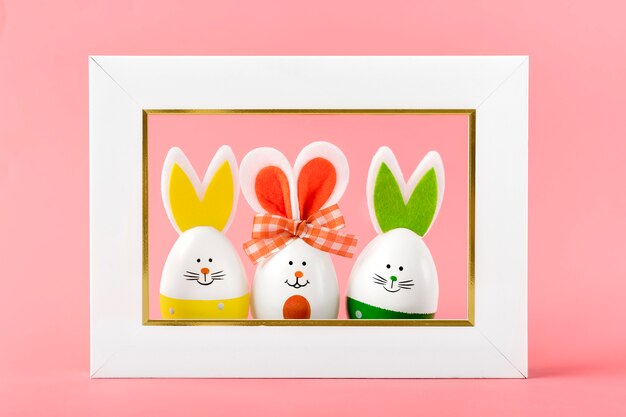 Painted easter eggs cute bunny with smile faces, ears, bow and white photoframe on coral pink backgroung.