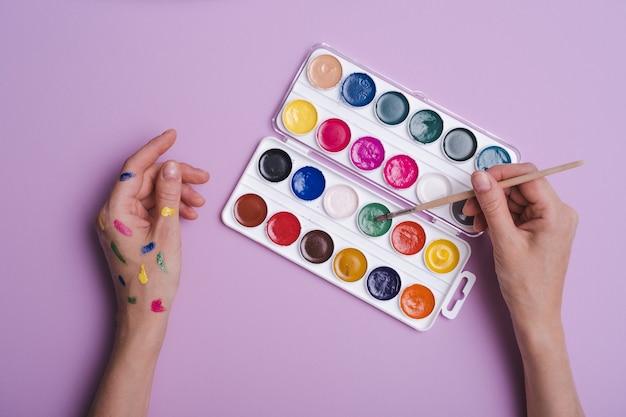 Mani dipinte con pallet di colori ad acquerello e pennello su sfondo viola, pensiero creativo, nuovo concetto di creatività, vista dall'alto