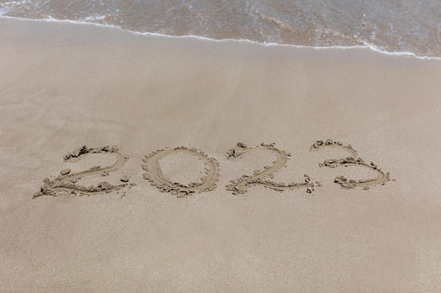 Painted new year numbers 2023 on the sand in the summer at sea which will soon be washed away by the approaching wave