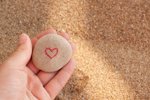 Раскрашенное красное сердце на плоской гальке в руке на фоне песка с местом для текста