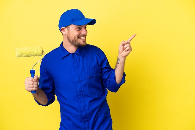 Painter Brazilian man isolated on yellow background pointing finger to the side and presenting a product