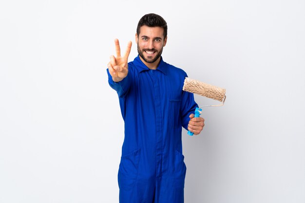 Painter man holding a paint roller isolated on white wall smiling and showing victory sign