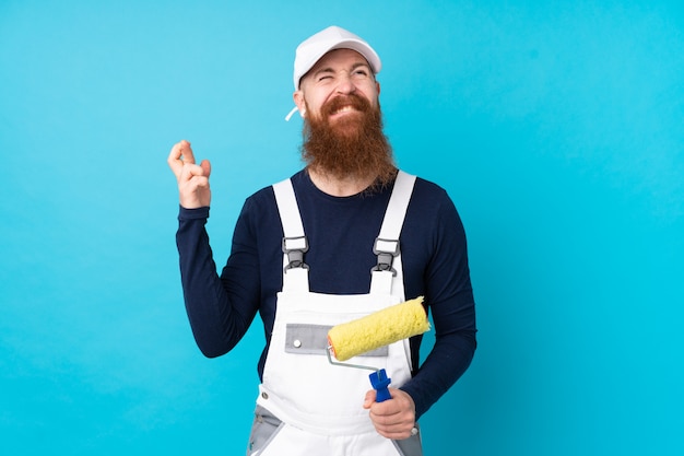 Uomo del pittore con la barba lunga sopra la parete blu isolata con l'incrocio delle dita