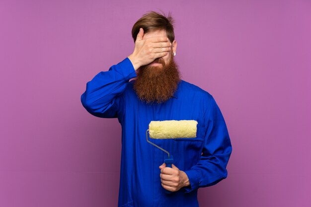 Painter man with long beard over isolated purple covering eyes by hands