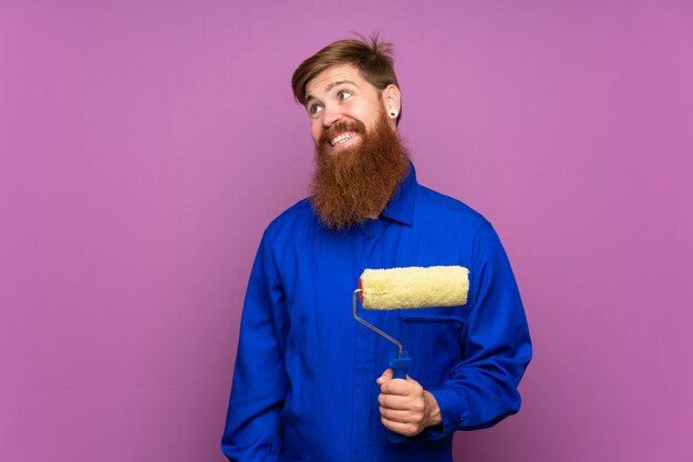 Painter man with long beard over isolated purple wall laughing and looking up