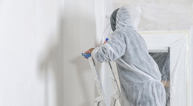 painter stands with paint roller on a ladder to paint the room window with white color. do it yourself concept image