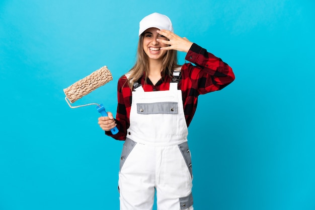 Painter woman over blue wall covering eyes by hands and smiling