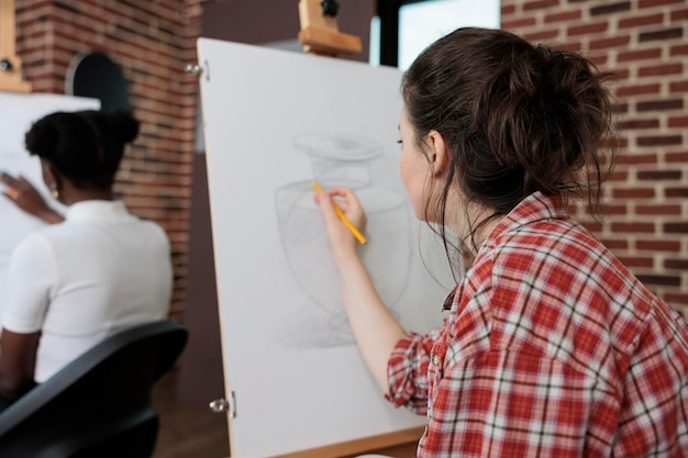 Painter woman drawing artistic vase on white canvas working at illustration technique using graphic pencil during art lesson in creativity studio. Multiethnic team developing creative skill