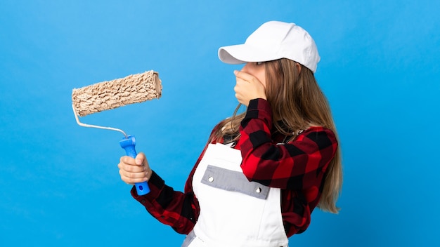 Painter woman over isolated blue wall covering mouth and looking to the side