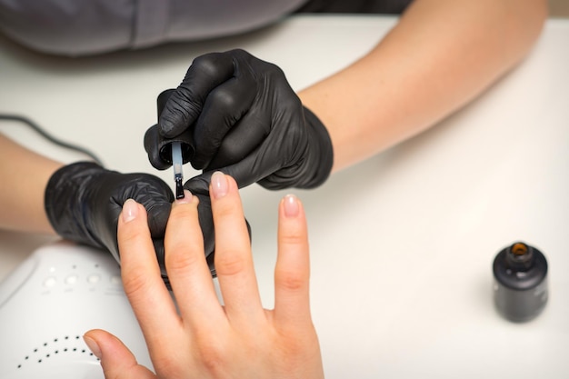 Painting nails of a woman. Hands of Manicurist in black gloves applying transparent nail polish on female Nails in a beauty salon.