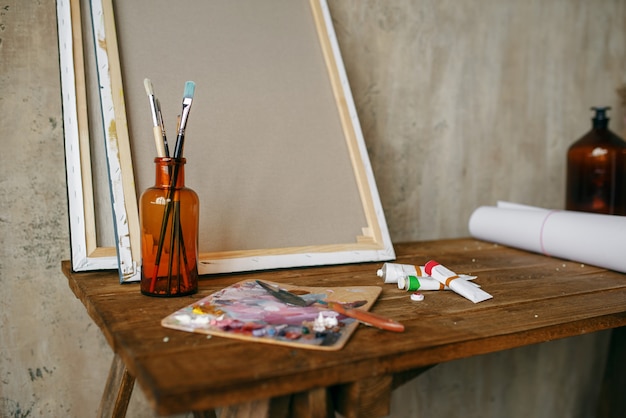 paints on palette, brush in bottle, canvas, nobody. painter tools on the table in art studio, equipment on artist workplace, paintbrush, creative atelier or workshop