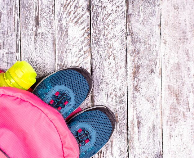 ピンクのバックパックと白い木の表面の水のためのスポーツボトルの青い女性のスニーカーのペア