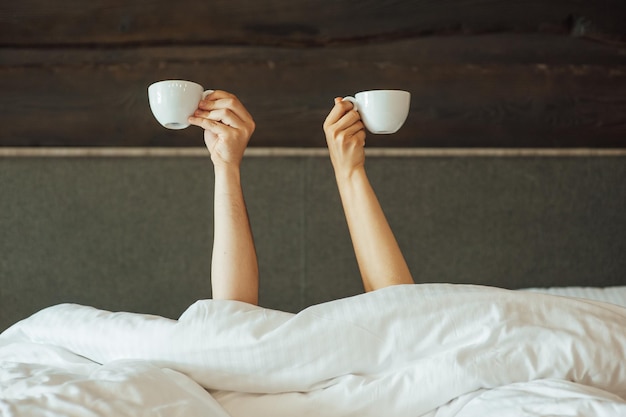 A pair of hands are holding over the blanket in the bed are\
holding cups of coffee. morning time.