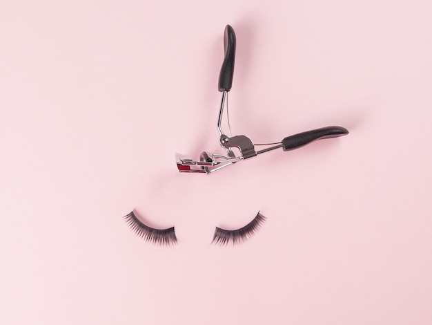 A pair of large false eyelashes and metal eyelash curlers on a pink background