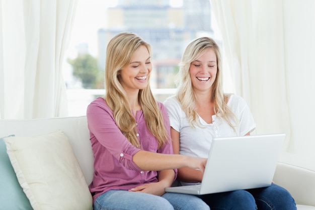 A pair of laughing women sitting on the couch with a laptop in hand