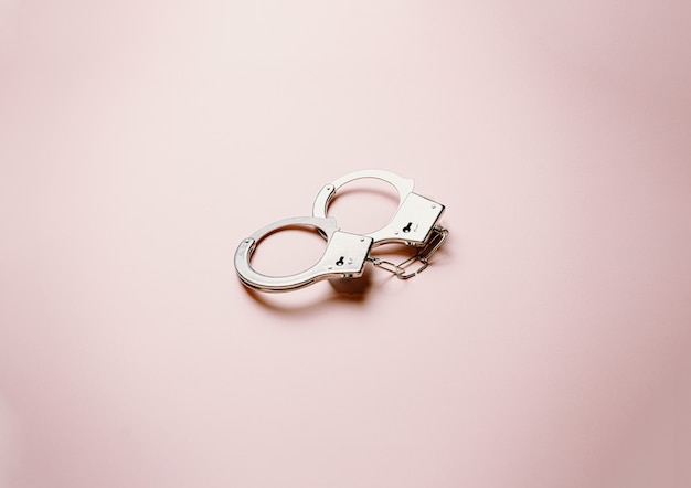 A pair of metallic handcuffs over a flat and bright pastel pink background