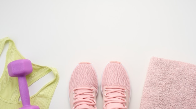 A pair of pink sneakers, a terry towel and a plastic dumbbell on a white background, top view, copy space