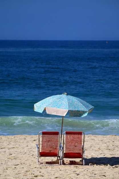 Пара красных шезлонгов и синий зонтик на песчаном пляже против голубого океана