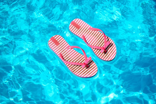 Pair of red and white striped flip flops floating on top of the blue water in a sparkling sunlit swimming pool in summer