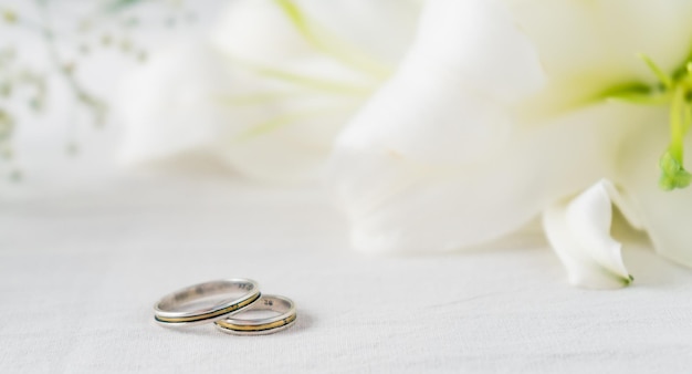 A Pair of wedding rings on a white surface with beautiful white lilies flowers in the background. Commitment and love concept. Copy space.