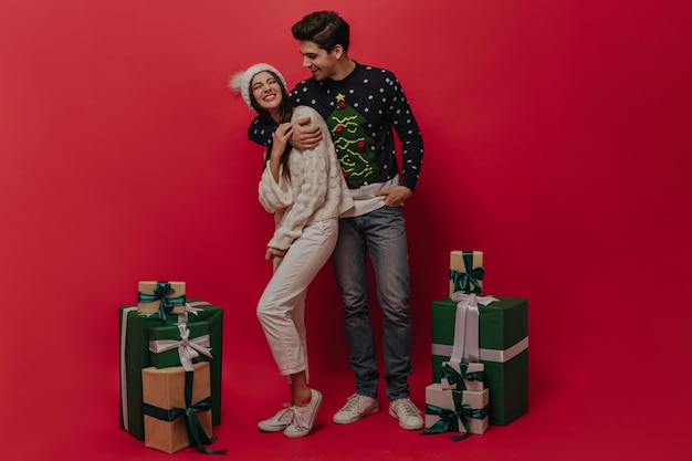 Pair of young brunettehaired people in cozy winter outfits having fun and posing near many gift boxes against plain red background