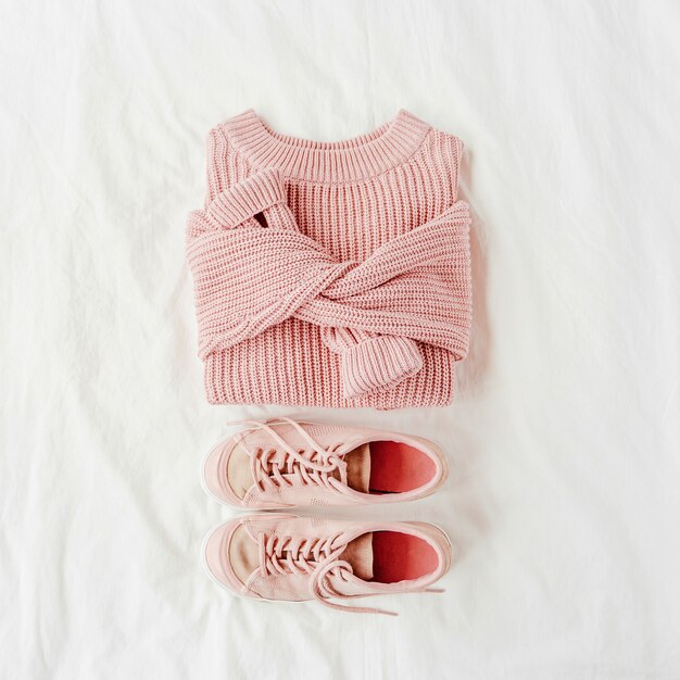 ベッドの上の淡いピンクの暖かいセーターとスニーカー。女性のスタイリッシュな秋冬の服。居心地の良い冬のルック。フラットレイ、上面図。