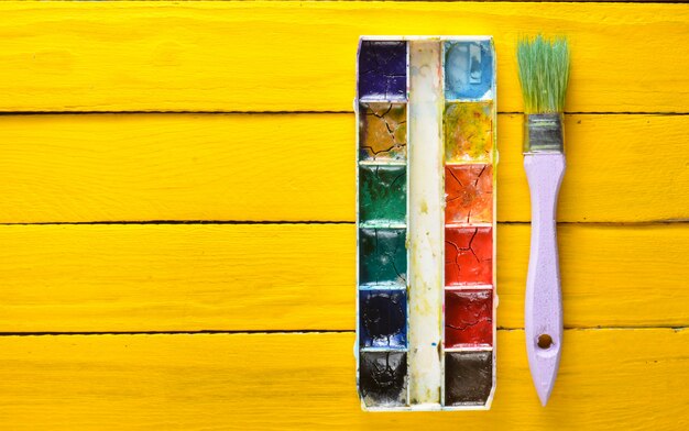 A palette of watercolor paint and a brush for painting on a yellow wooden boards. Top view. Copy space. Art concept.
