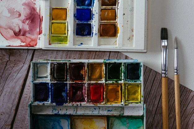 Palette of watercolor paints blank paper and brushes on wooden background Artist's work space