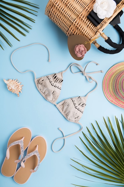 Palm leaves, fashion hat, bikini, flip flops, straw beach bag on a light pastel blue surface, travel and vacations concept