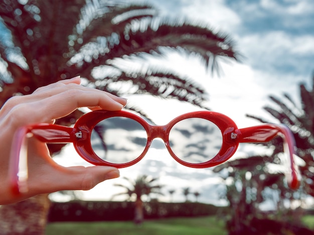 Пальмы, солнцезащитные очки. концепция отдыха. девушка держит в руке модные красные очки и готовится их надеть.