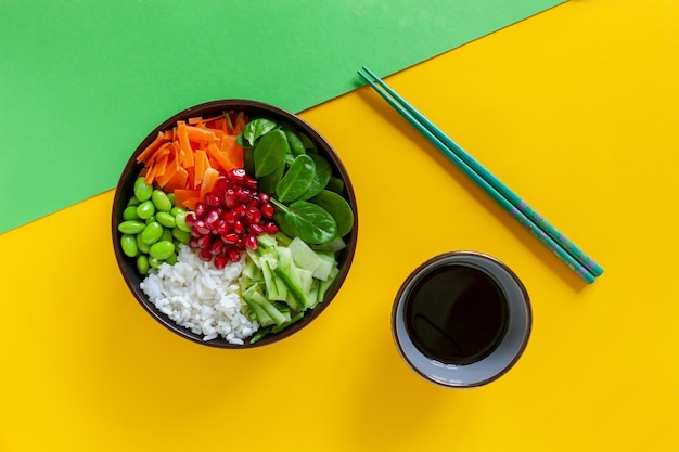 Pan Asian trendy Homemade Vegan Poke Bowl with Rice, edamame, fresh vegetables and soy sauce, served with chopsticks on vivid yellow and green background