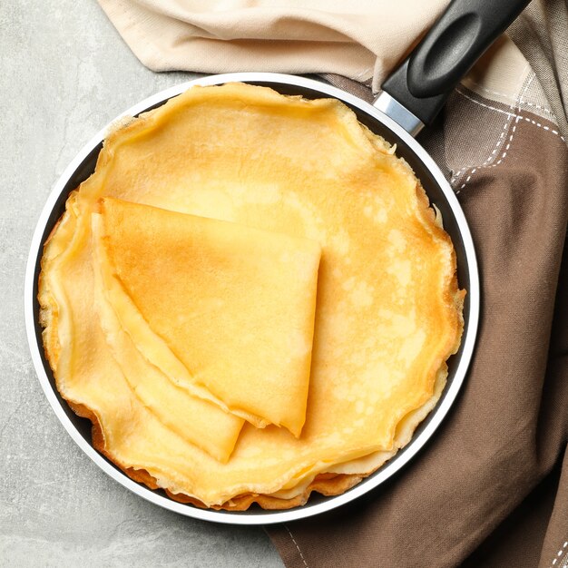 Pan with thin pancakes and kitchen towel on gray background