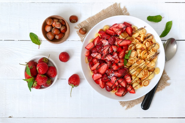 Pancake cereal in a bowl with fresh strawberries and nuts on a white wooden surface