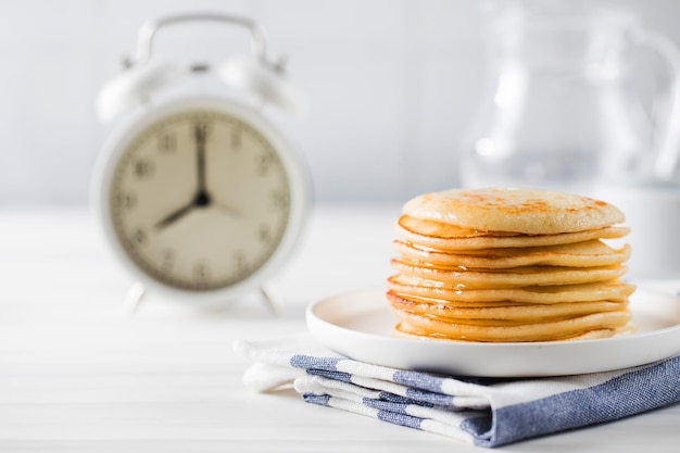 The pancakes are stacked in a plate In the background a jug of milk and an alarm clock Breakfast concept