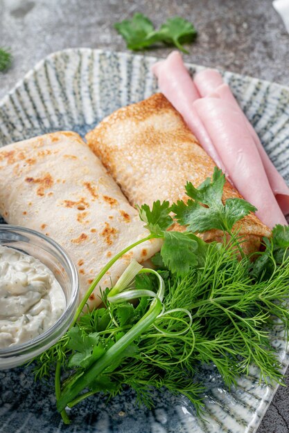 Pancakes filled with ham in a plate with parsley and dill greens and sour cream on a gray stone table, flatlay