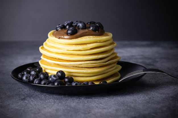 Pancakes pile with blueberries and chocolate paste on black plate with fork on gray table closeup