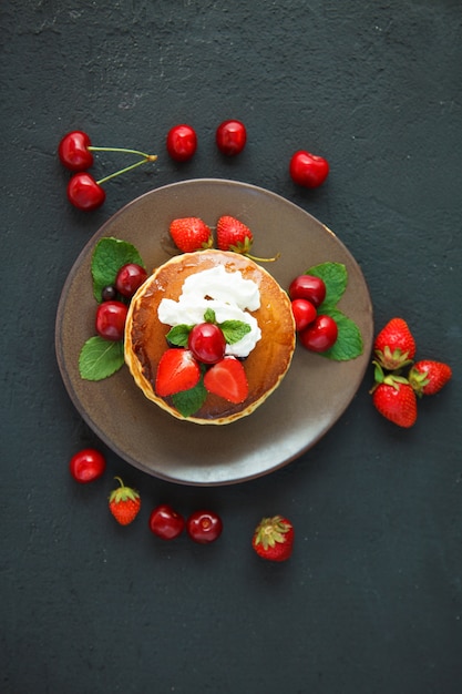 Pancakes in a plate with strawberries, whipped cream, mint, honey and cherry on a dark black background.