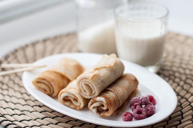 Pancakes rolled up on a white plate with frozen raspberries on a wicker napkin