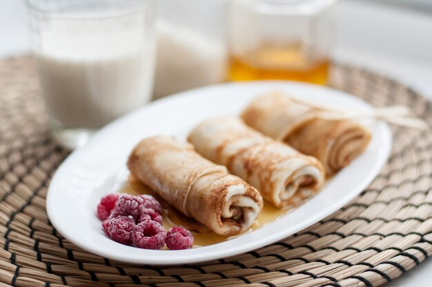Pancakes in rolls on a white plate with frozen raspberries and honey on the background of milk, cans of sugar and honey