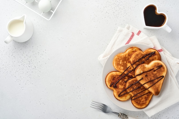 灰色のセラミックプレートにチョコレートソース、灰色のコンクリートの背景にコーヒーのカップと朝食の心の形をしたパンケーキ。バレンタインデーの朝食のテーブルセッティング。上面図のコピースペース。