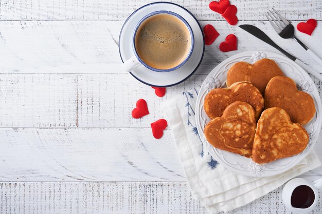 Frittelle con marmellata di frutti di bosco e miele a forma di cuore e tazza di caffè calda su sfondo bianco di legno. colazione concettuale per san valentino o piacevole sorpresa per la persona amata tavolo visto dall'alto