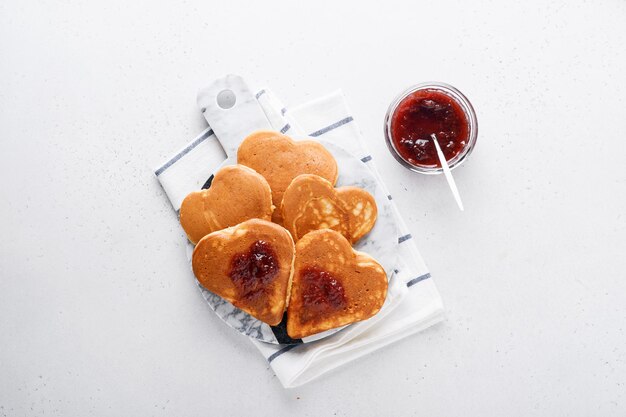 Pancakes with berry jam and honey in shape of heart and hot cup of coffee over white wooden background. Concept breakfast for Valentines Day or pleasant surprise for loved one Table viewed from above