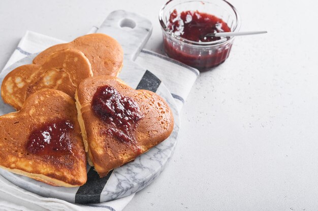 Pancakes with berry jam and honey in shape of heart and hot cup of coffee over white wooden background Concept breakfast for Valentines Day or pleasant surprise for loved one Table viewed from above