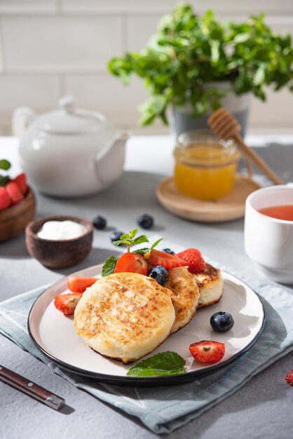 Pancakes with cottage cheese or syrniki with fresh berries, honey and sour cream on a blurred white brick background with a cup of tea. The concept of a healthy and delicious breakfast.