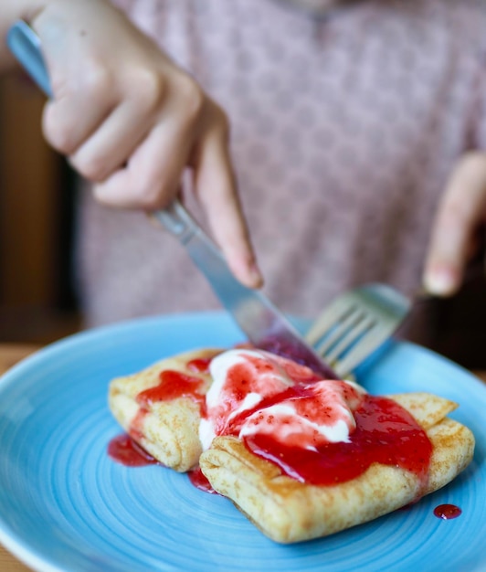 青い皿にジャムとサワークリームを乗せたパンケーキを人のクローズアップが食べる