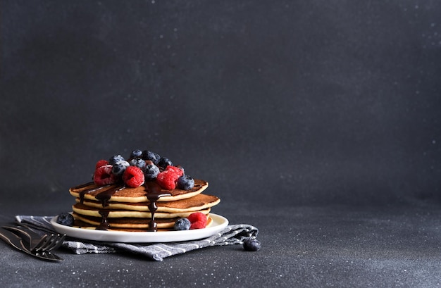 Pancakes with raspberries and blueberries and chocolate sauce on a dark concrete background Stack of pancakes with berries