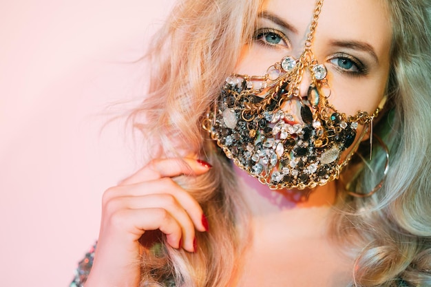 Pandemic beauty. DIY accessory. Glamour carnival look. Portrait of sensual blonde woman in creative handmade gold chain gem face mask on pink neon light background.