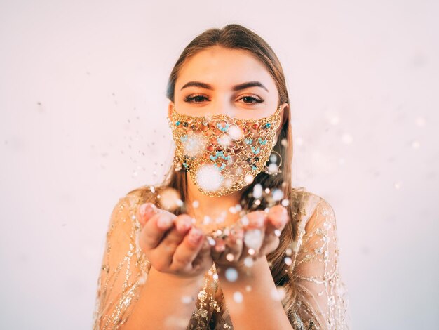 Pandemic Christmas. Festive greeting. Quarantine party celebration. Amused woman in glamour gold chain face mask catching bokeh light snowflakes in open palms on pastel pink copy space background.