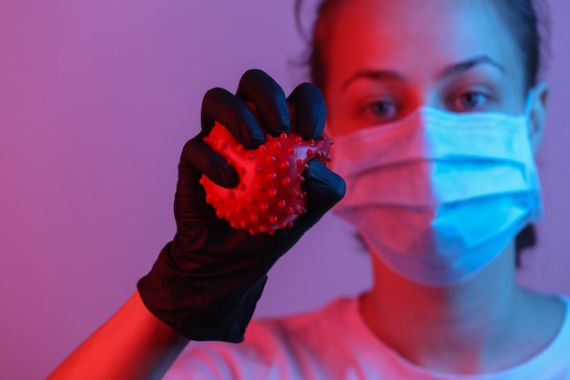 Pandemic covid-19 theme. Woman in protective gloves, medical face mask holds a virus strain model. Red blue gradient neon light