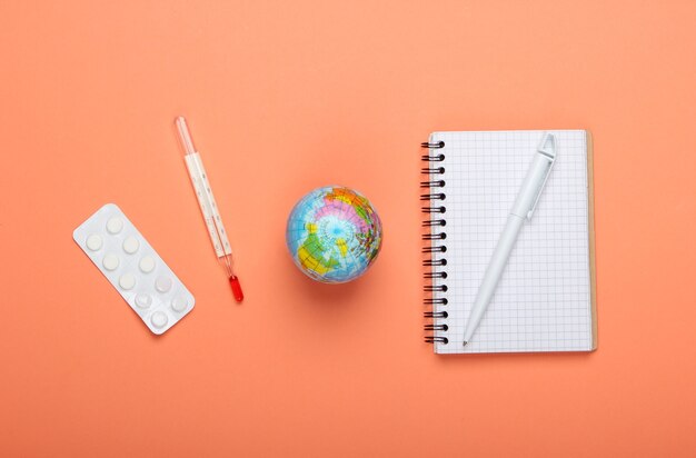 Pandemic outbreak or global warming concept. Globe, thermometer, notebook and blister of pills on orange background. Top view